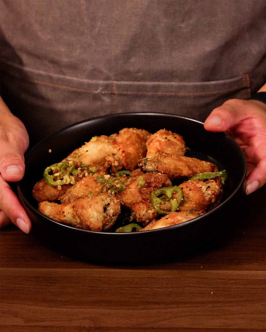 A person in a gray apron holding a dark bowl of crispy air crisper salt and pepper chicken wings topped with sliced green chilies, minced garlic, and scallions on a wooden surface.