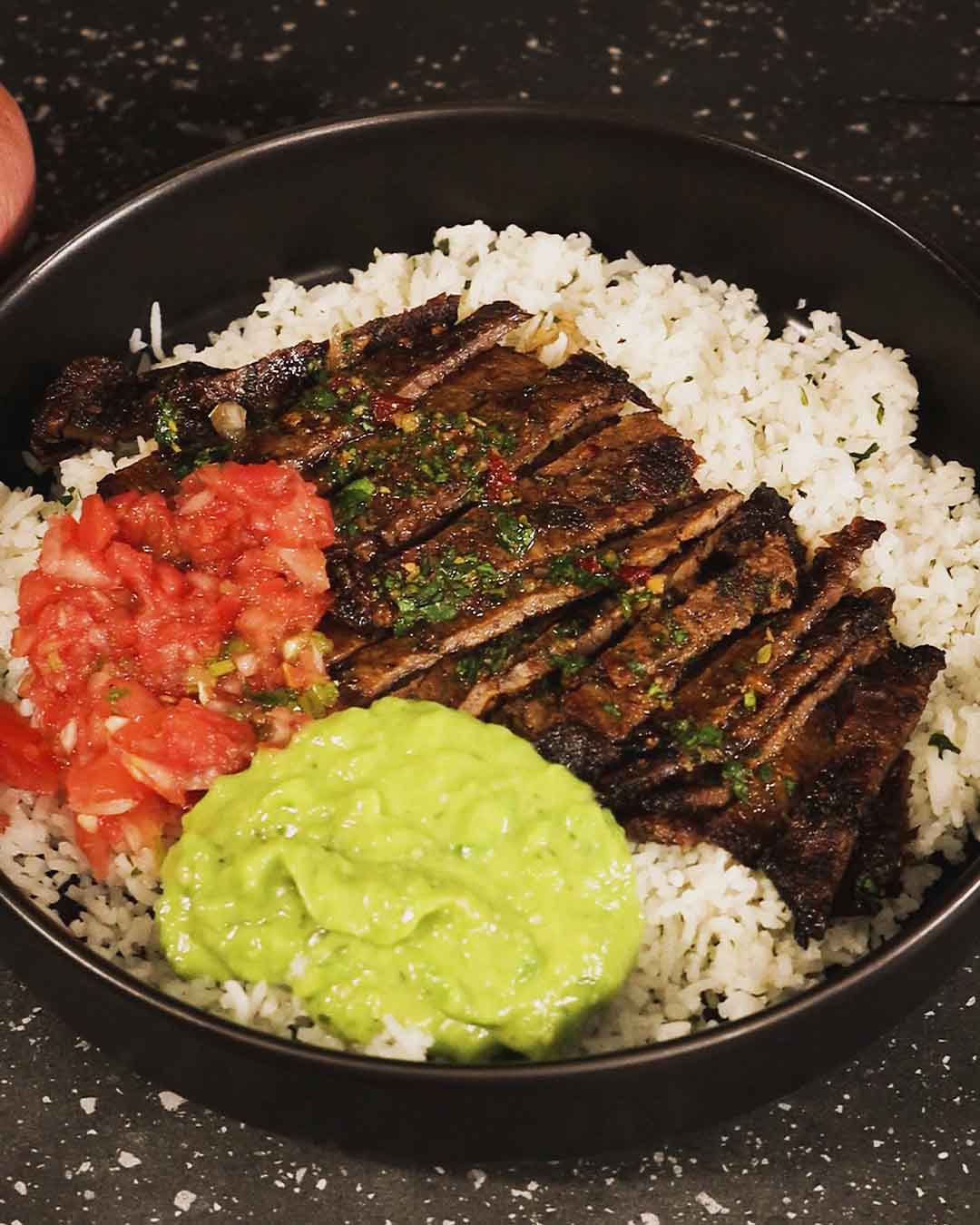 Black bowl with sliced chimichurri ribeye steak over white rice, guacamole, and fresh tomato salsa.