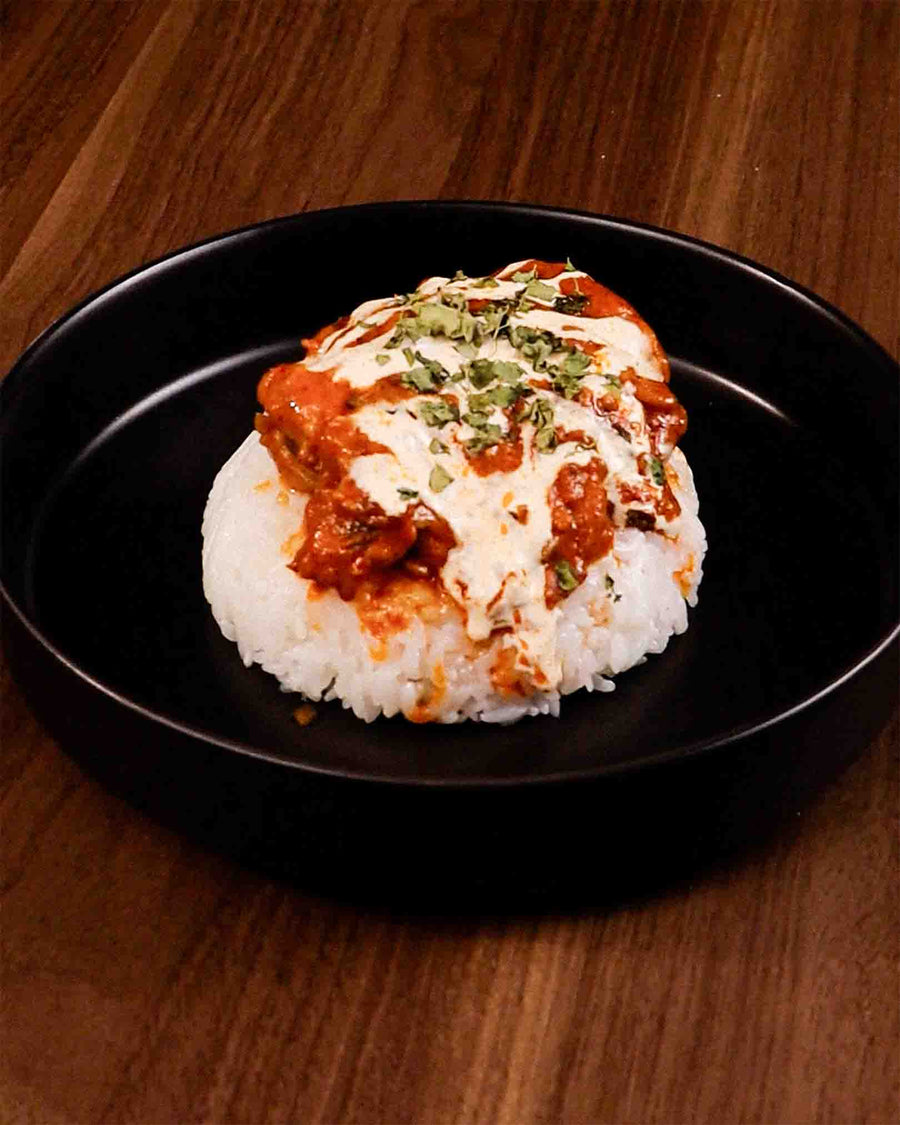 Butter chicken with steamed rice, creamy tomato sauce, and herb garnish in a black bowl.